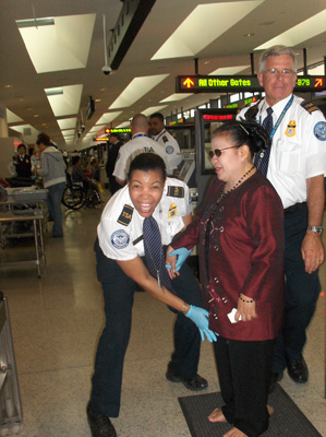 Nanta is standing and smiling while a woman with a security uniform holds a wand in front of her, grinning at the camera. Behind Nanta is another security guard, smiling.